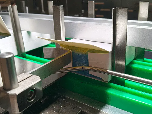 the-major-flaps-are-folded-closed-over-the-pre-folded-minor-flaps-by-stationary-folding-rails-positioned-along-the-sides-of-the-conveyor-path